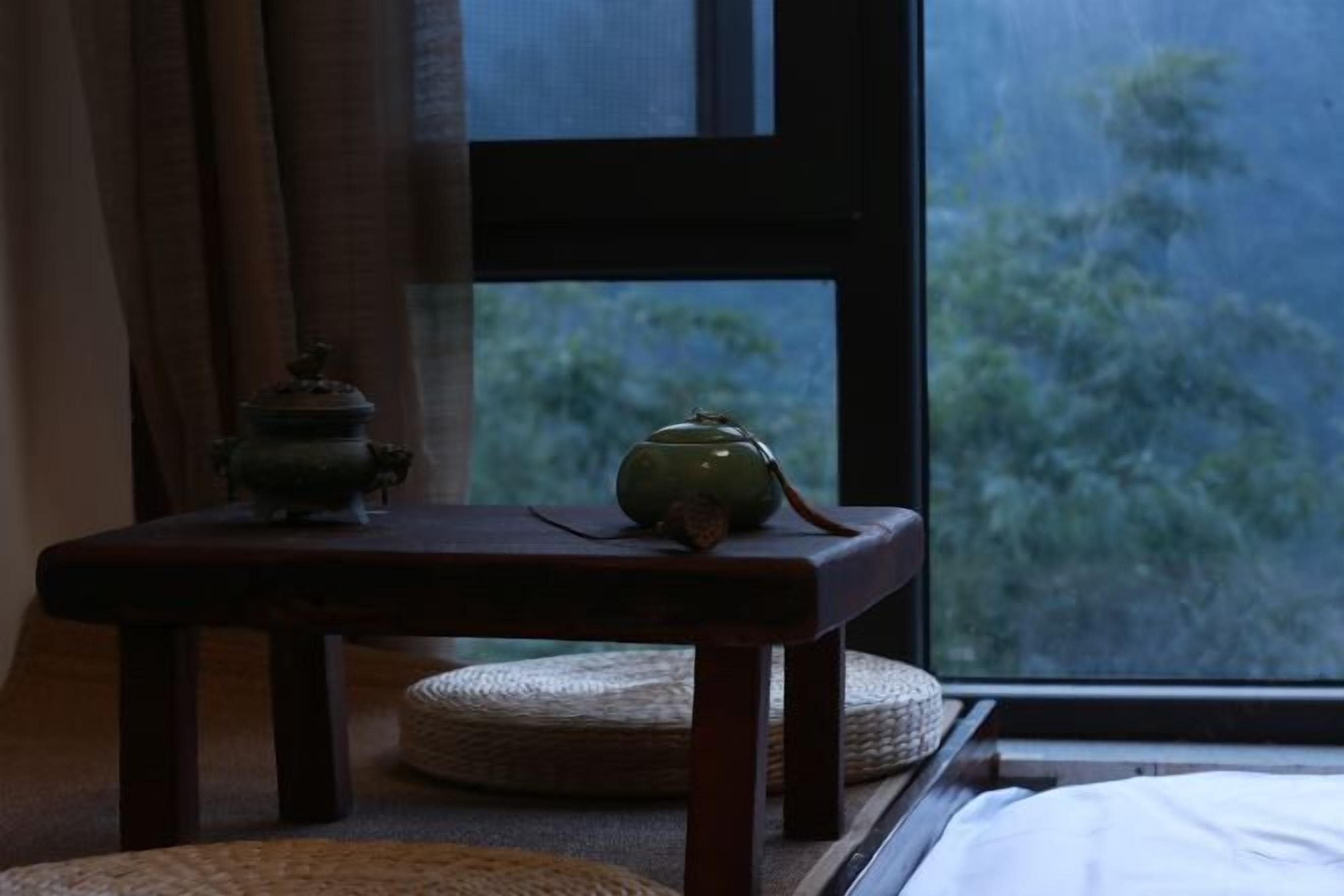 A room with a bed, a window with a view of trees, a small table with a teapot, and a lamp.