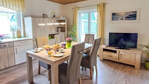 Uma sala de jantar com mesa de madeira, cadeiras e uma televisão. Há uma janela com cortinas, uma planta e um quadro na parede.