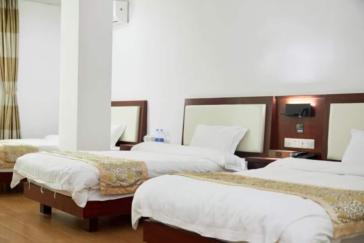 A hotel room with three beds, each with white bedding and a decorative headboard. There is a nightstand with a lamp and a small table with a bottle of water.