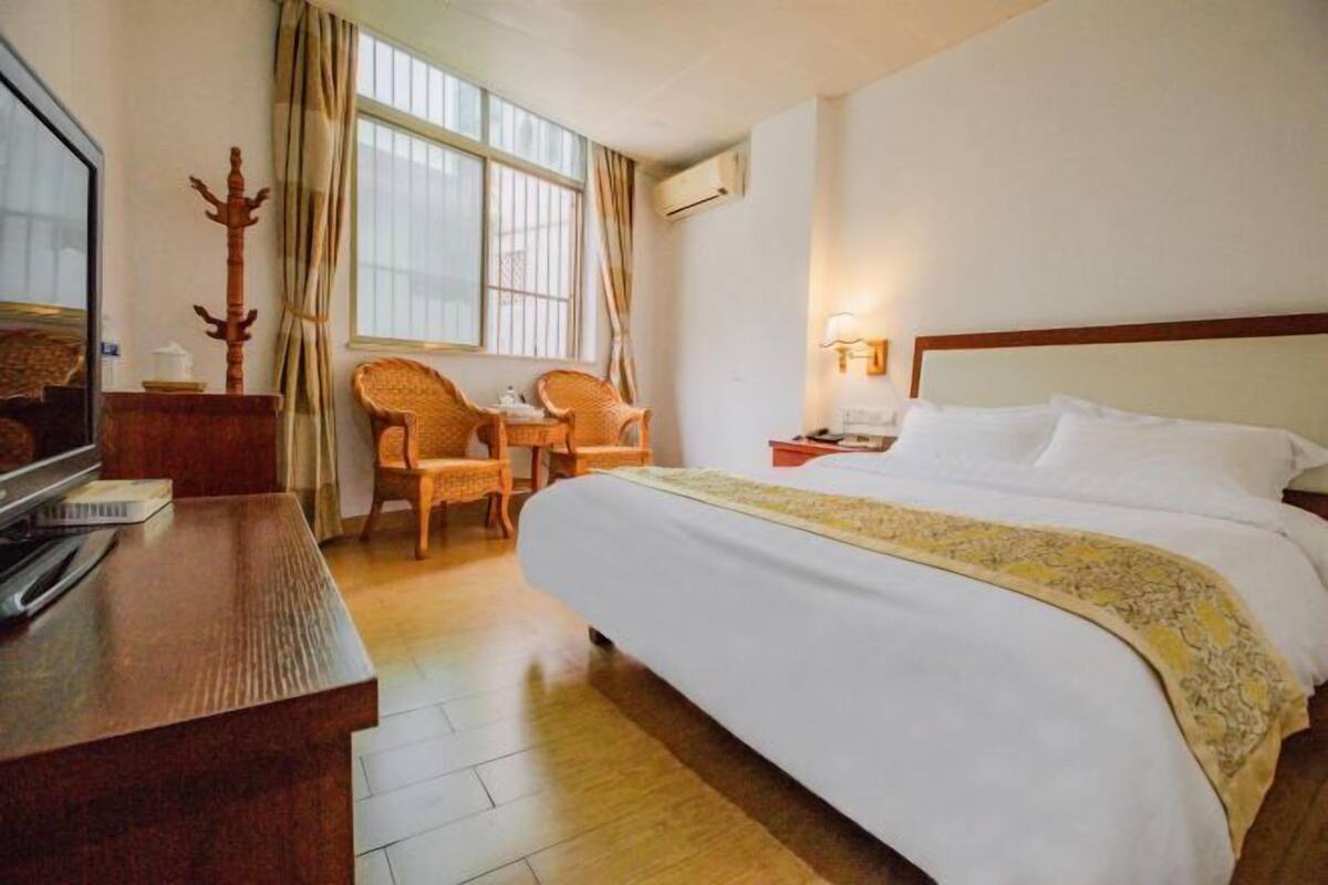 A hotel room with a bed, two chairs, a desk, a TV, and a window with curtains.