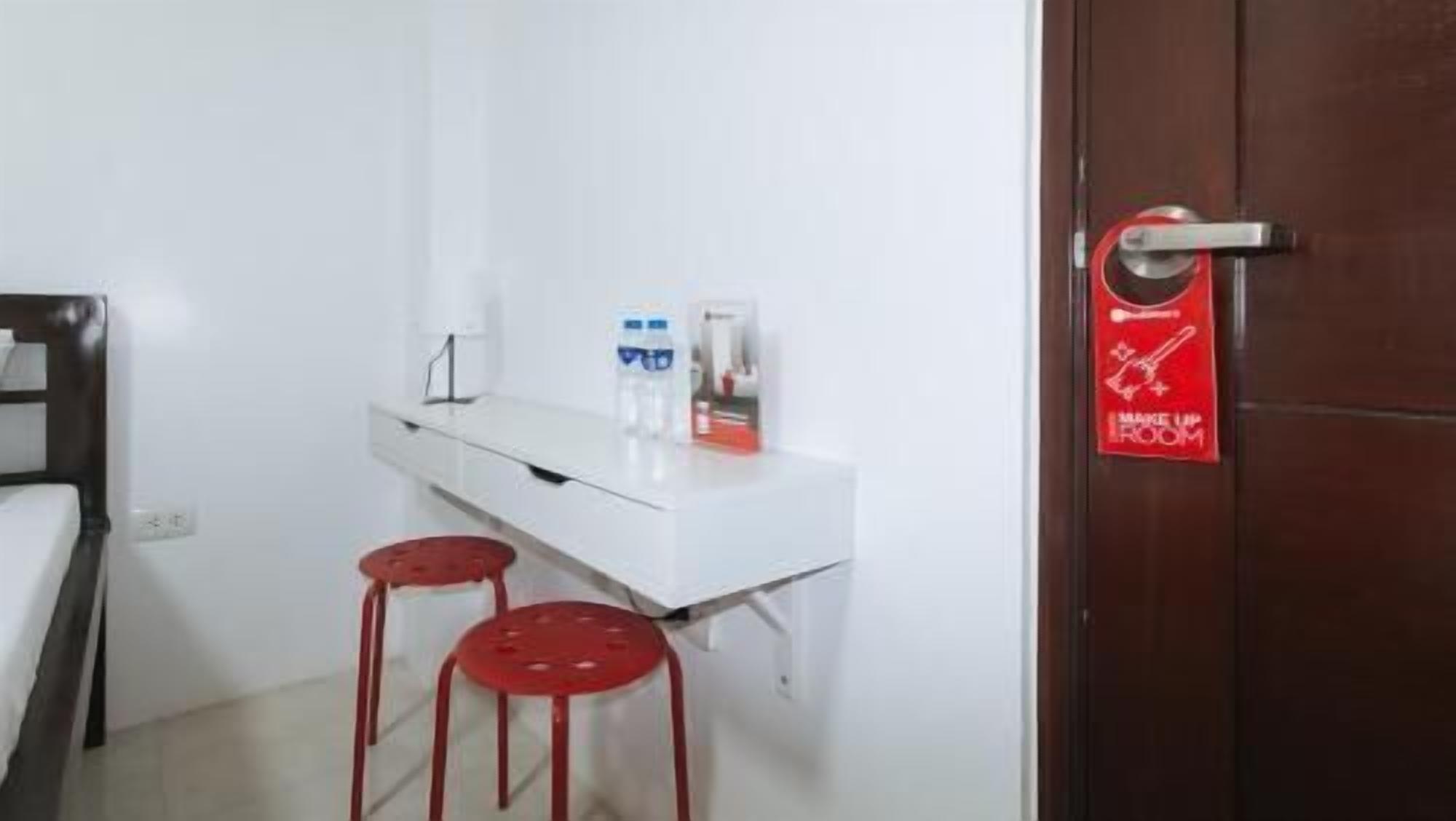 A small room with a white desk, two red stools, a white wall, and a door with a red sign.