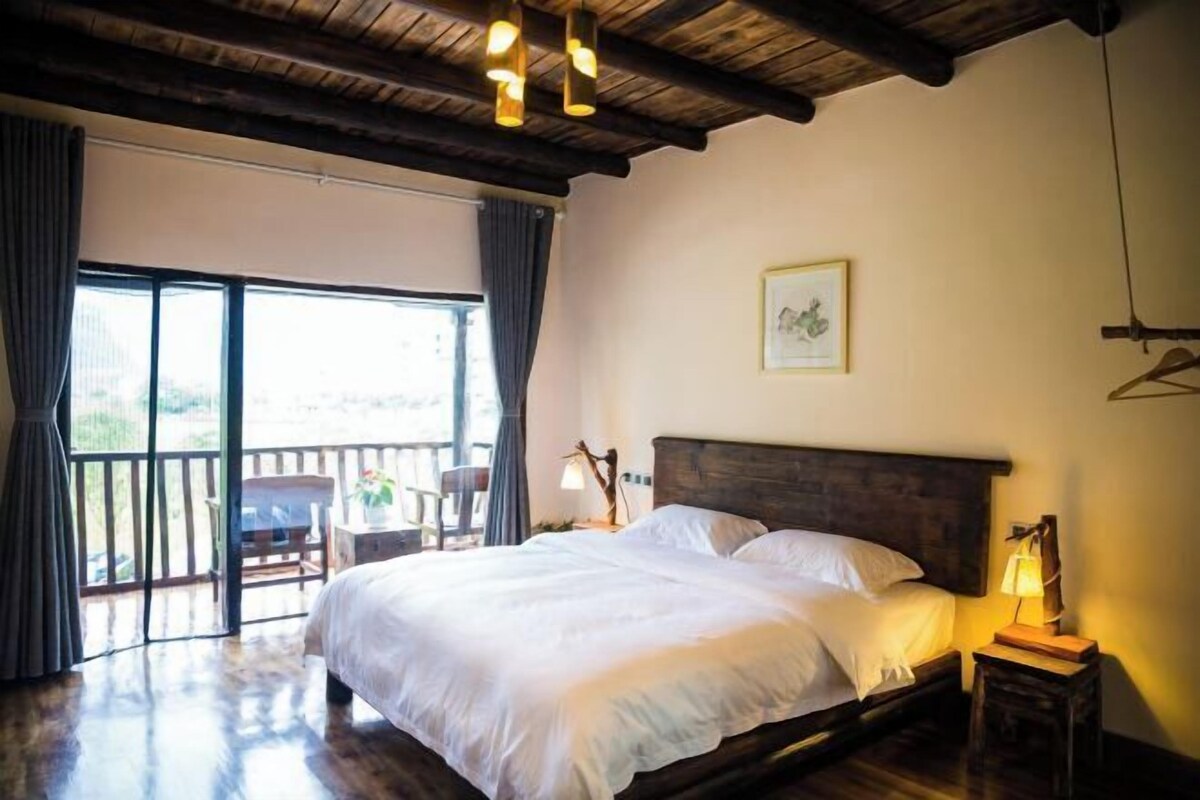 Ein Schlafzimmer mit einem großen Bett, Holzbalken an der Decke, einem Balkon mit einem Sessel und einem gerahmten Bild an der Wand.