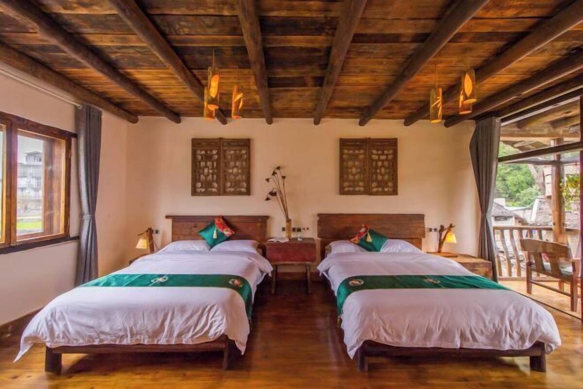 A room with two beds, wooden beams on the ceiling, a window with curtains, and a view of greenery outside.