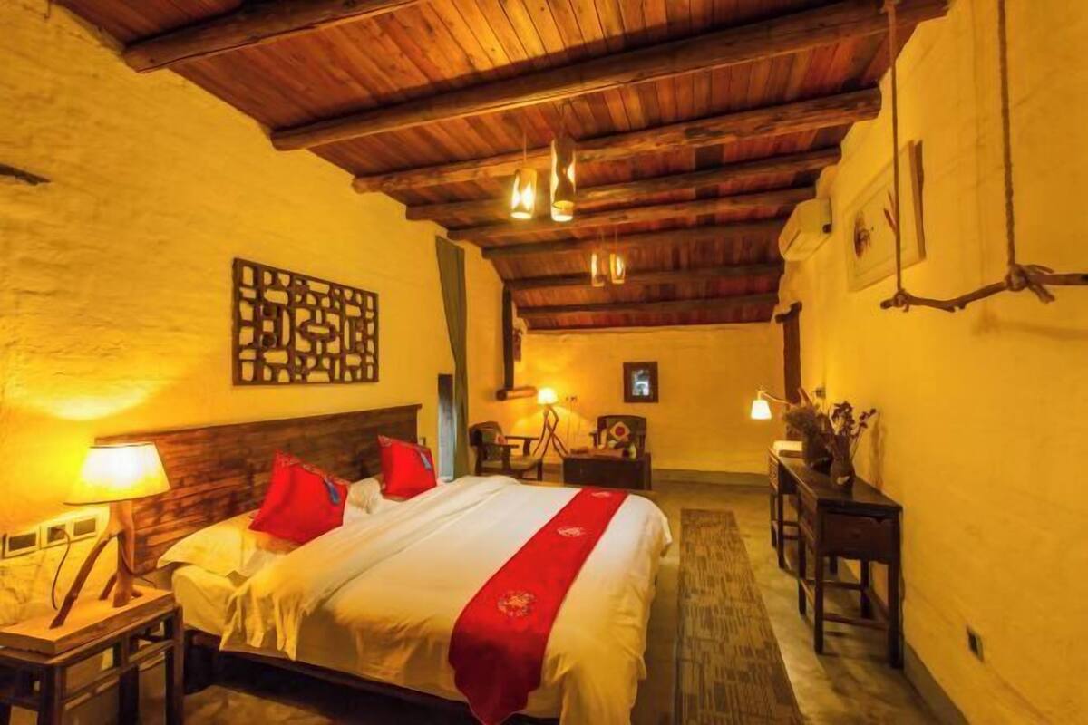 A hotel room with a wooden ceiling, a bed with red pillows, a nightstand with a lamp, a desk with a chair, and a wall-mounted picture.