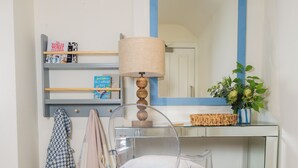 Une salle de bain avec un fauteuil transparent, un miroir doté d’un cadre bleu, une lampe et une étagère avec des livres et une plante.