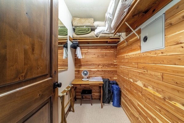 Una habitación pequeña de madera con un escritorio, una silla y un taburete. Hay un ventilador sobre el escritorio, y mantas dobladas y un colchón en la pared.