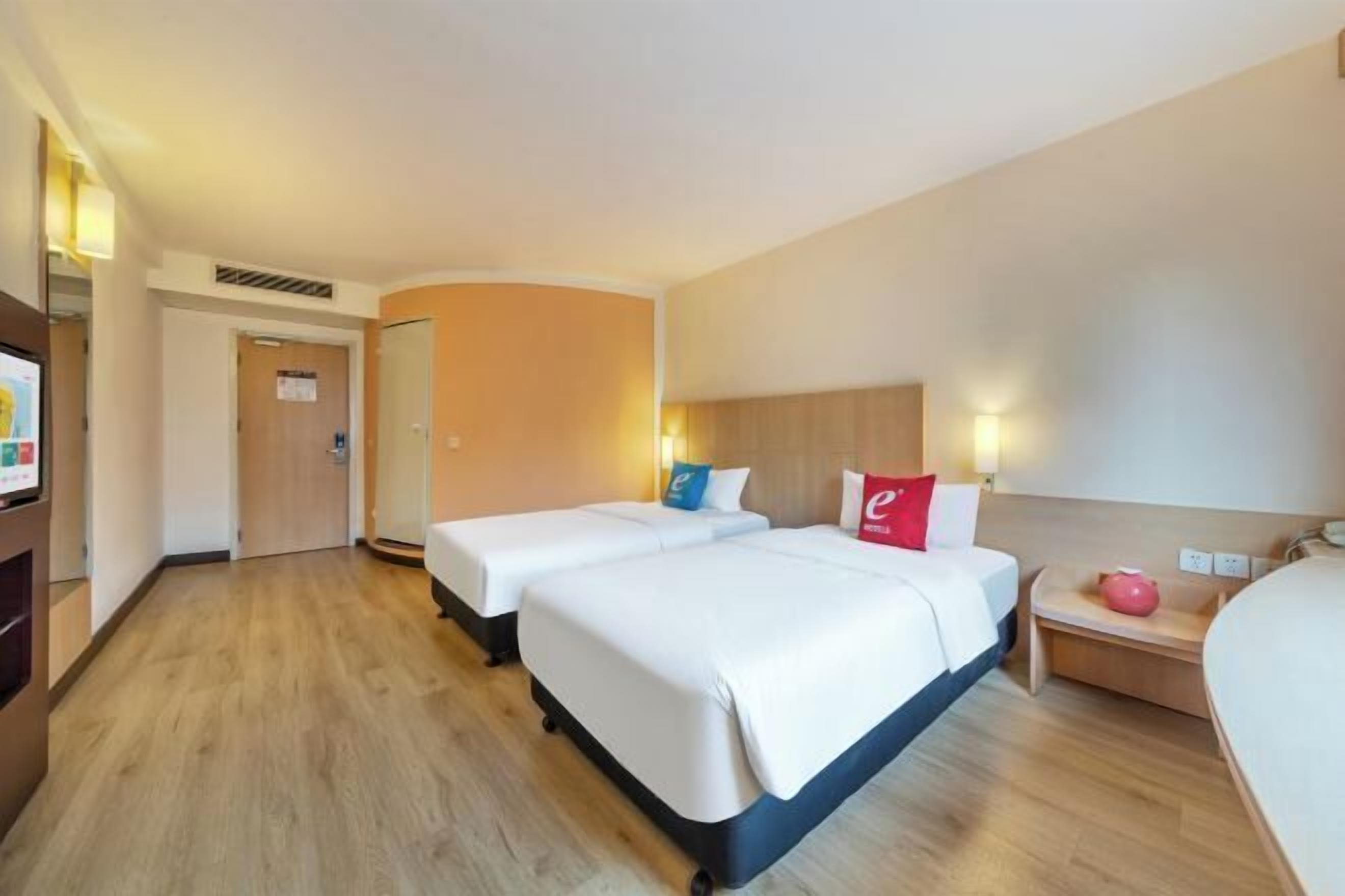Habitación de hotel con dos camas individuales, un escritorio, una silla, un televisor y una pequeña mesa con un objeto rosa.