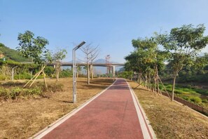 Un chemin pavé à la surface rouge, bordé d’arbres et d’arbustes, qui mène vers un pont situé au loin.
