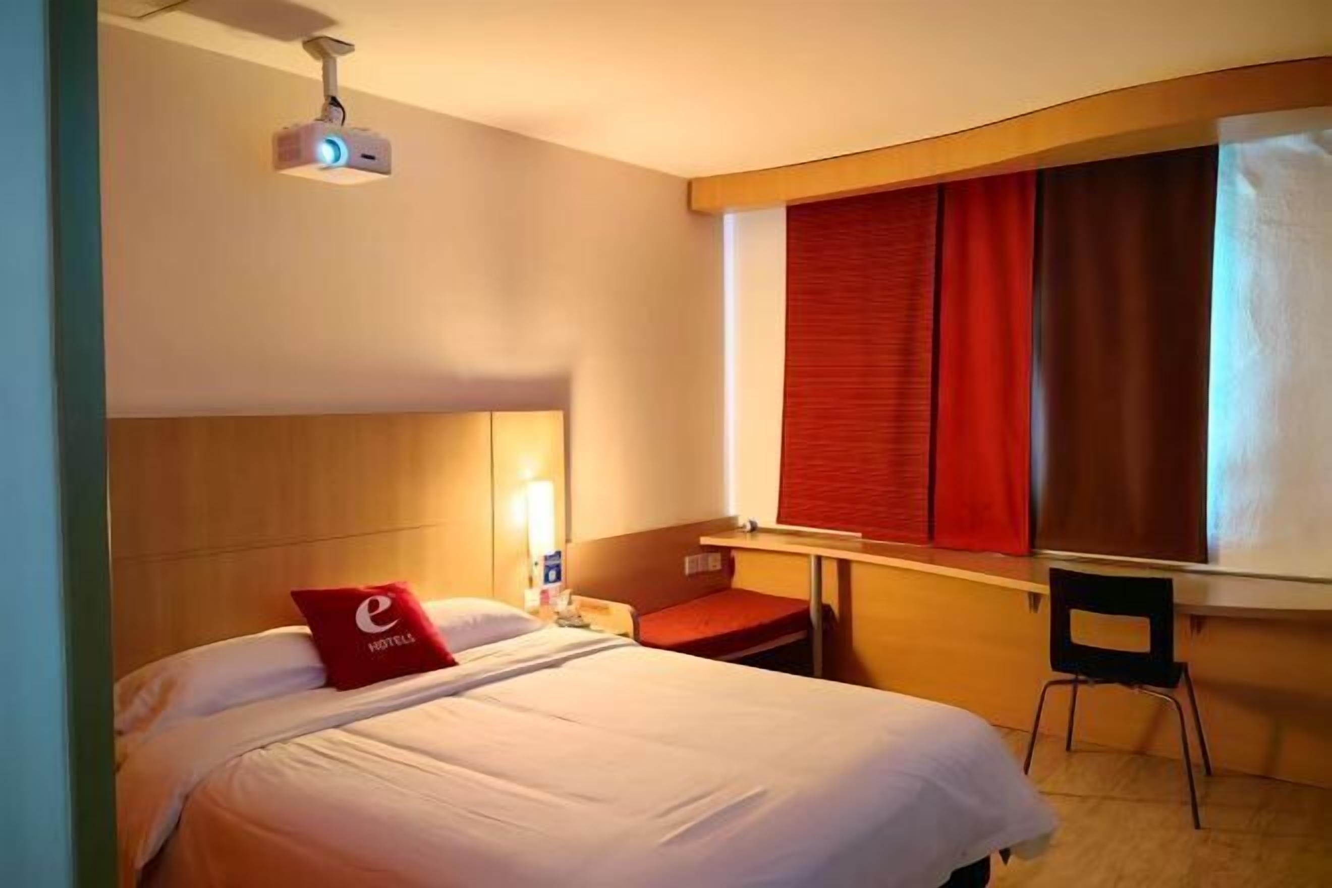 A hotel room with a bed, a desk, a chair, a projector, and a window with curtains.