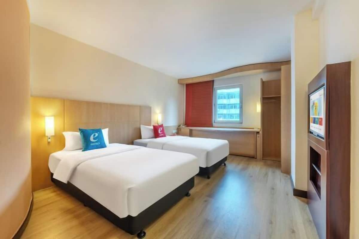 A hotel room with two beds, a desk, a TV, and a window with a view of buildings.