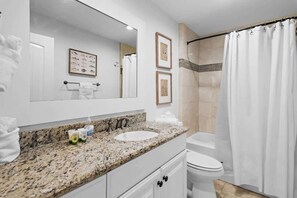 Une salle de bain avec un lavabo doté d’un plan de travail en granit, un grand miroir, des œuvres d’art encadrées, un rideau de douche et des toilettes.