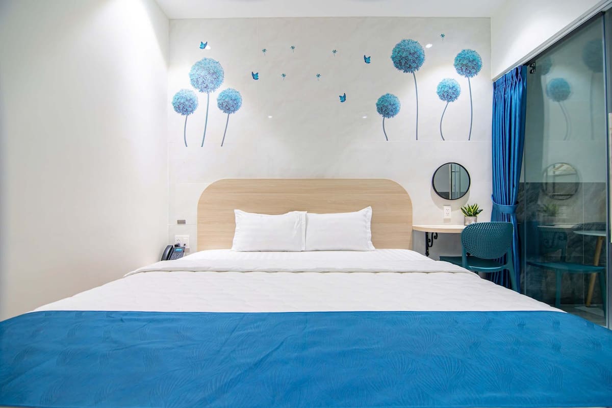 Ein modernes Hotelzimmer mit einem großen Bett, einem Nachttisch mit Telefon, einem kleinen Tisch mit Pflanze und einem blauen Vorhang an einer Glasscheibe.