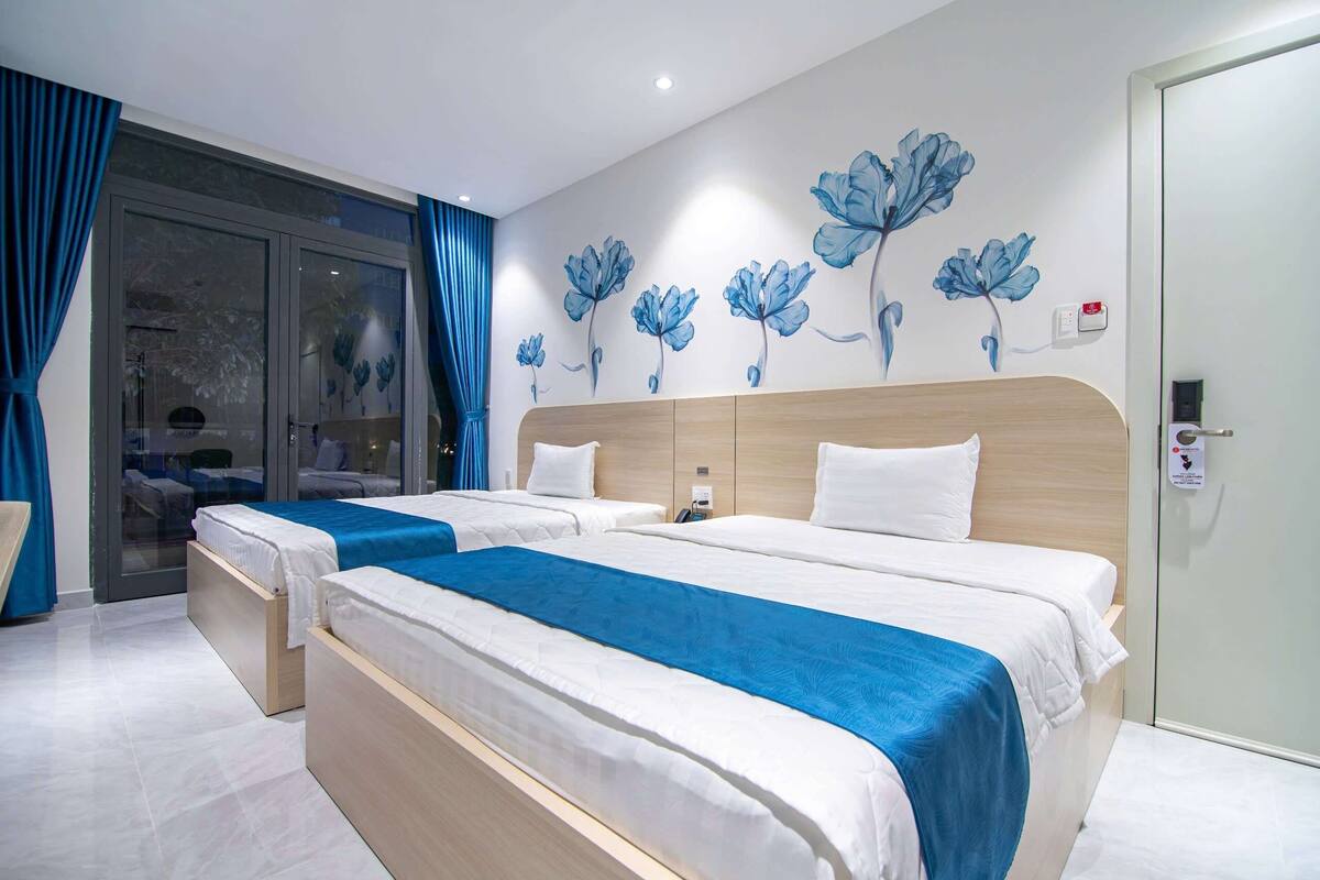 Ein Hotelzimmer mit zwei Betten, einem großen Fenster mit Vorhängen und einer Wandmalerei mit Blumenmotiven.