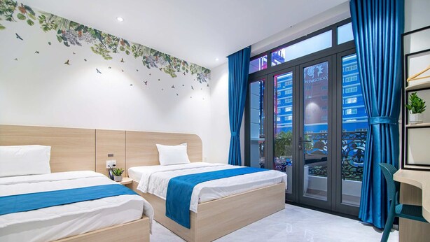 A modern hotel room with two beds, a large window with blue curtains, a city view, and a small desk with a chair. - Bin Bin Hotel 14 - Near Vstar School D7 (Ho Chi Minh City)