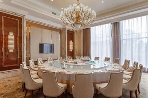 Ein Konferenzraum mit rundem Tisch, Stühlen, einer Kandelaberleuchte, einem Flachbildfernseher und großen Fenstern.