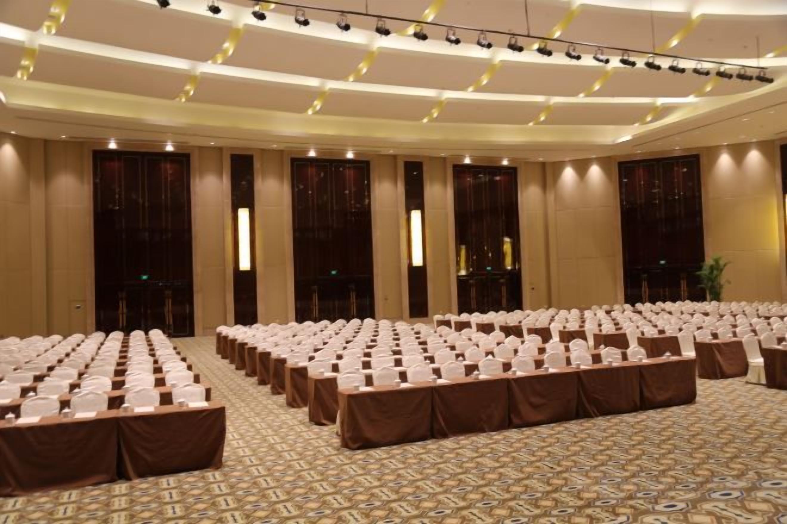 Een ruime congreszaal met rijen tafels en stoelen, een kamerbreed tapijt met patroon, en een hoge plafond met verlichtingsarmaturen.