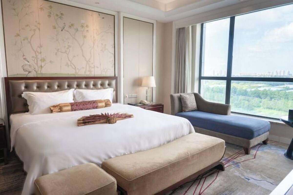Een hotelkamer met een groot bed, een bank, een leuning en uitzicht op de stad door grote ramen.