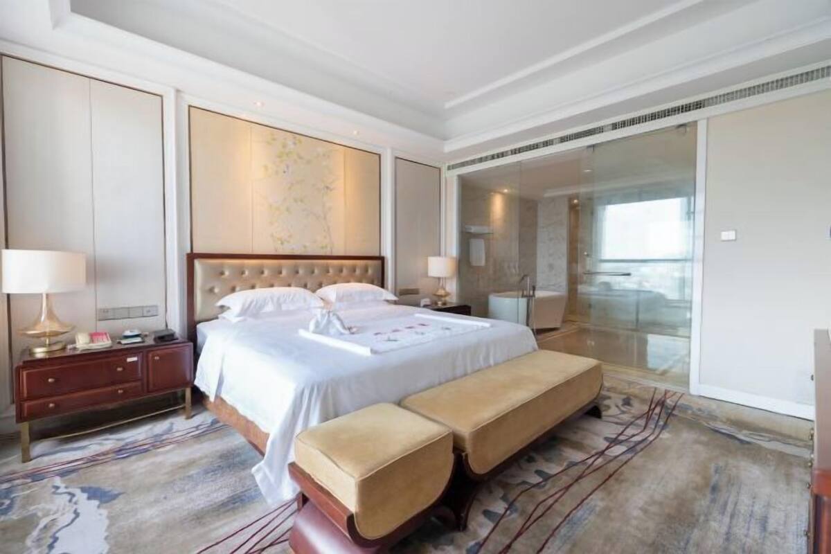 Hotelkamer met een groot bed, een bankje, een nachtkastje, een lamp, een glazen douchecabine en uitzicht op buiten.