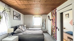 Un dormitorio con techo de madera, cama con ropa de cama gris, mesita de noche con lámpara, radiador y ventana con cortinas.