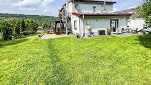 Una casa de dos pisos con una amplia escalera, un patio con mobiliario exterior y una vista de una colina boscosa al fondo.