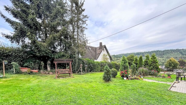 Ein gepflegter Garten mit einer Holz-Schaukel, einem weißen Haus mit Ziegeldach und verschiedenen Pflanzen und Bäumen.