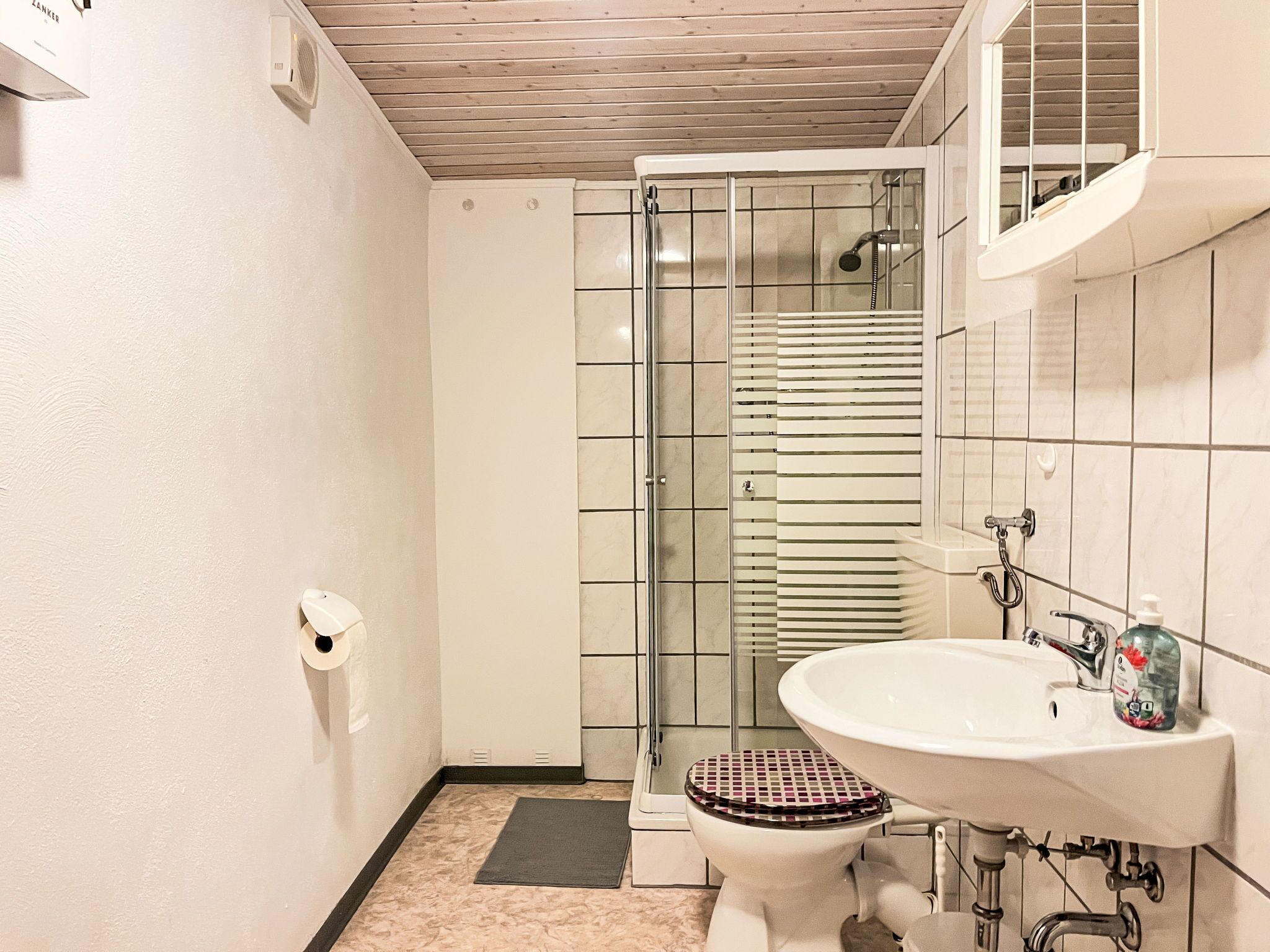 Een badkamer met een douchecabine van glas, een toilet met een decoratieve zitting en een wastafel met een zeepdispenser.