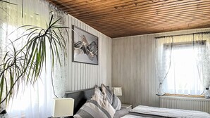 Un dormitorio con techo de madera, una cama con ropa de cama a rayas, una mesita de noche con lámpara y una planta grande en maceta.