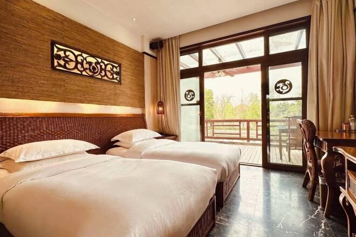A hotel room with two beds, a wooden headboard, a dining table, and a large glass door leading to an outdoor area.