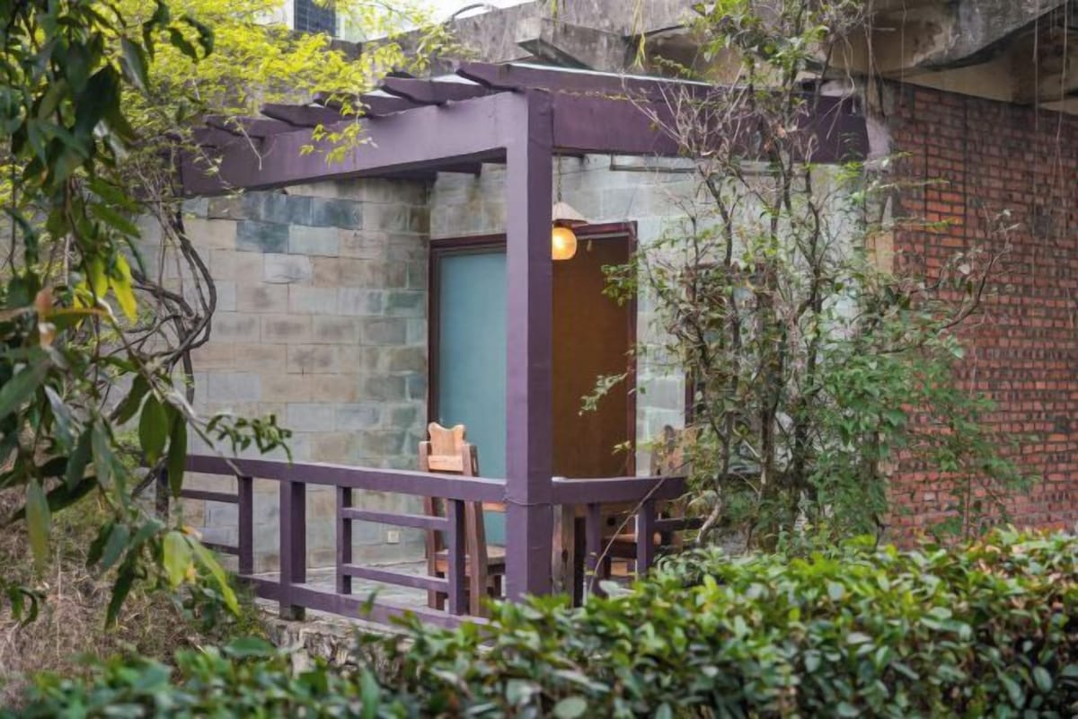 A small, enclosed outdoor space with a wooden bench and a door, surrounded by greenery and a brick wall.
