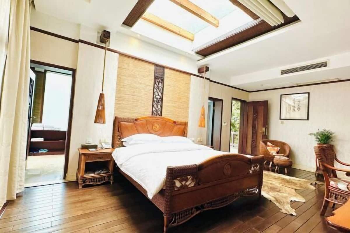 A bedroom with a large bed, wooden floor, a skylight, a nightstand, a chair, and a wall-mounted lamp.