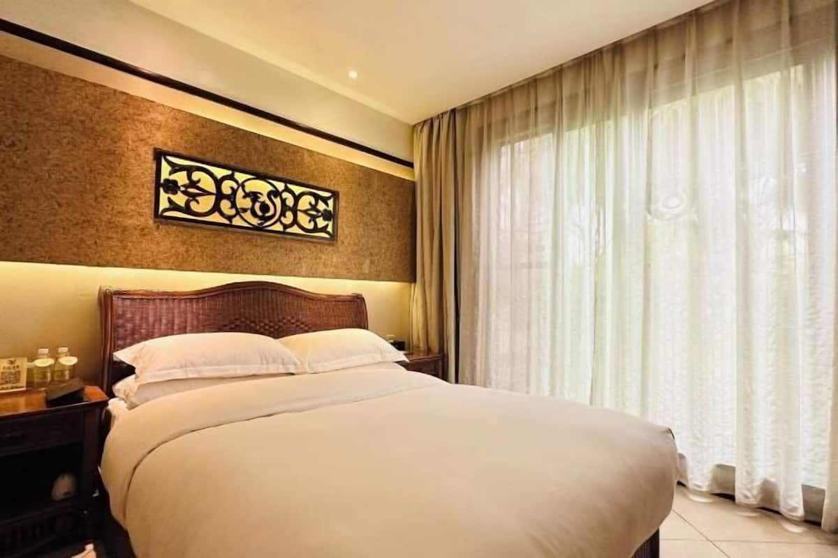 A hotel room with a large bed, a nightstand with a lamp, a wall art piece, and a window with curtains.