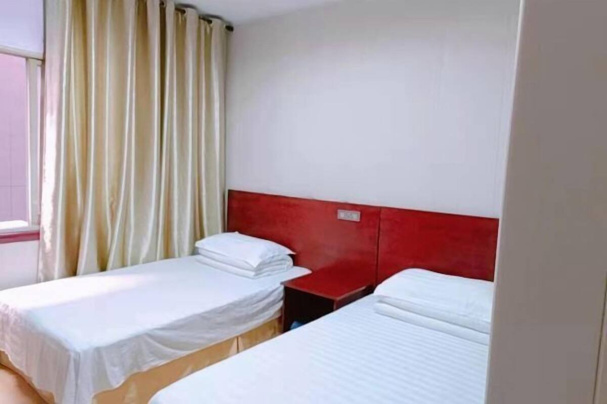 Een hotelkamer met twee enkele bedden, een rode hoofdbord, een raam met gordijnen en een witte muur.