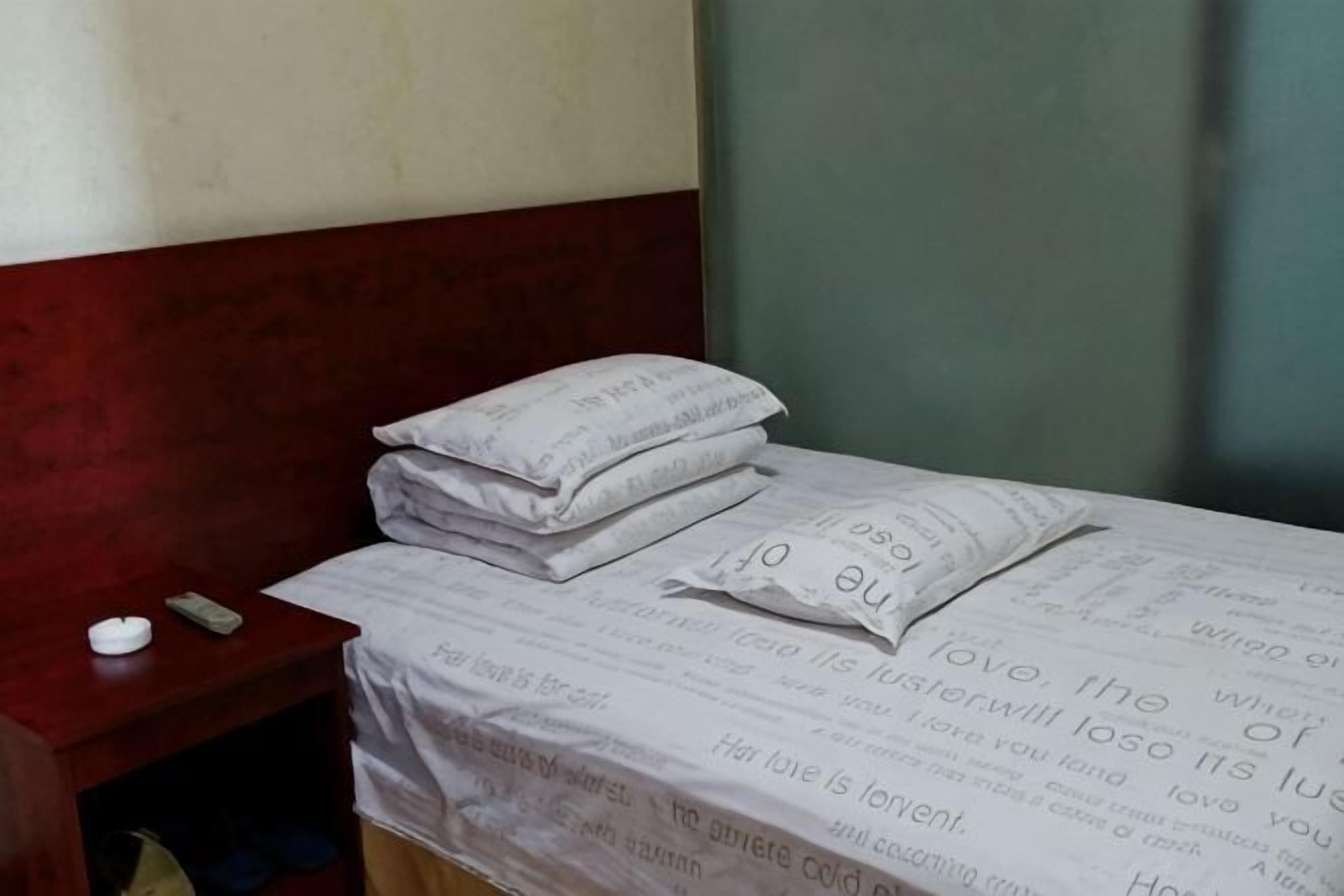 A bed with a white sheet and pillows, a bedside table with a remote control, and a plain wall.