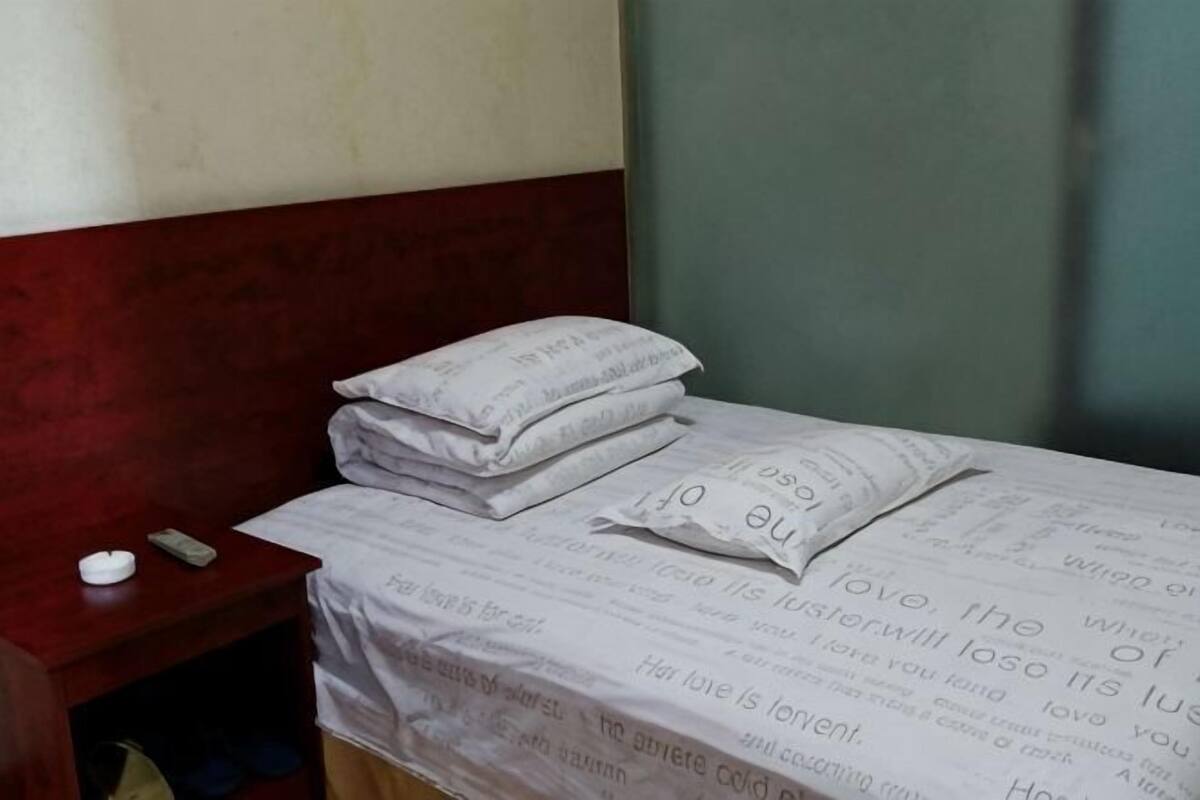 Een bed met een wit laken en kussens, een nachtkastje met een afstandsbediening en een effen muur.