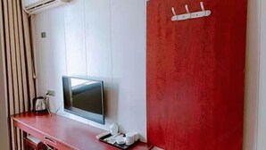 Ένα δωμάτιο με τηλεόραση επίπεδης οθόνης, έναν κόκκινο τοίχο που λειτουργεί ως διακοσμητικό στοιχείο, ένα ξύλινο γραφείο με βραστήρα και ένα κλιματιστικό τοποθετημένο στον τοίχο.