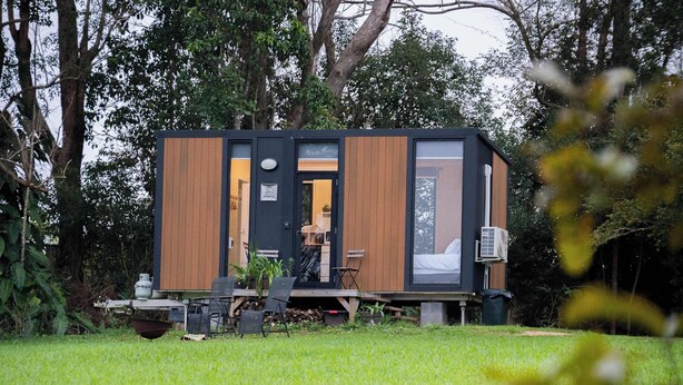 A modern, compact cabin with a wooden exterior, a visible door, and a small deck area with outdoor seating.