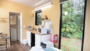 A compact hotel room with a bed, a small kitchenette, a mirror, and a window offering a view of greenery.