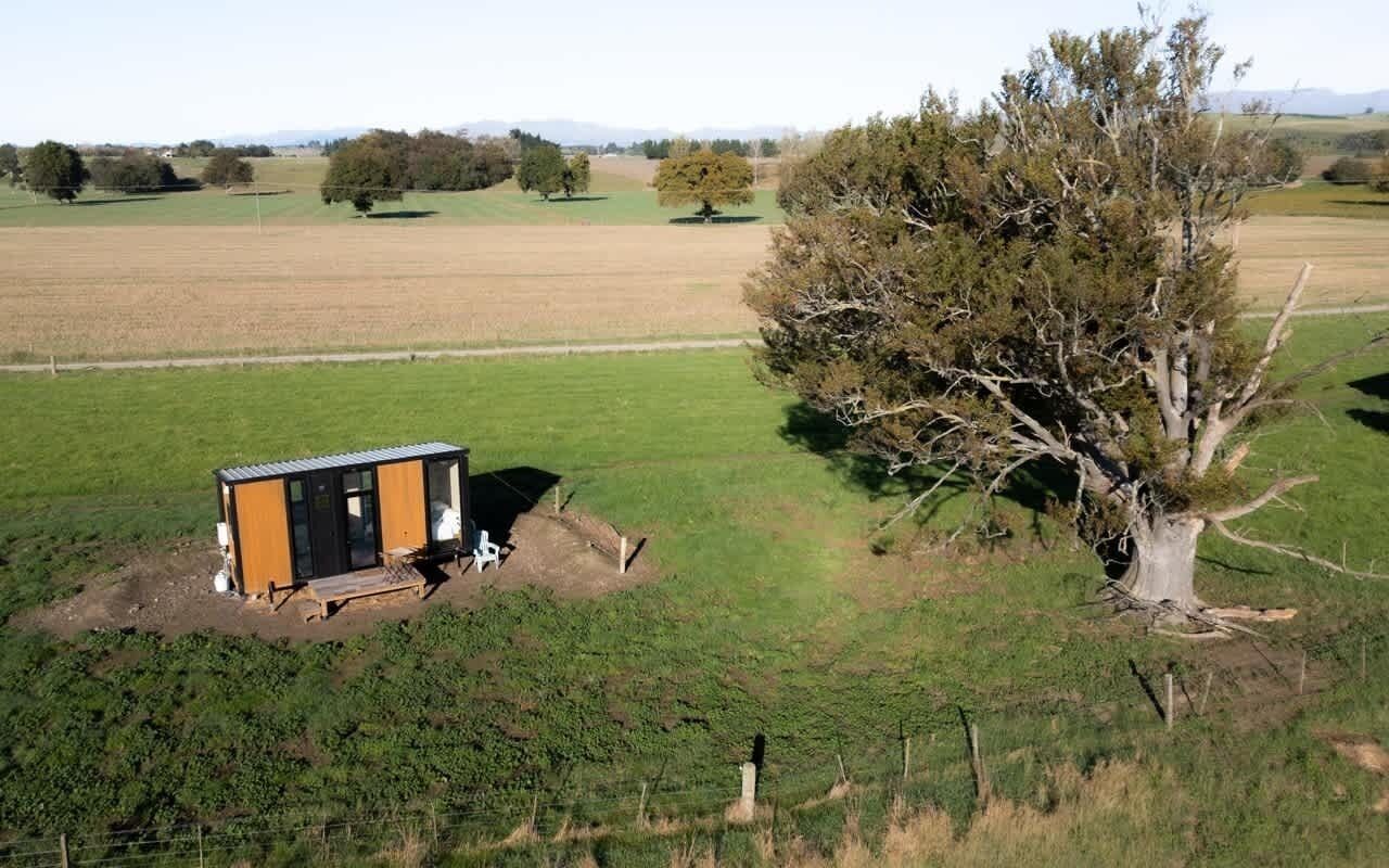 A small cabin with a metal roof and a wooden deck, surrounded by a grassy field and a large tree.