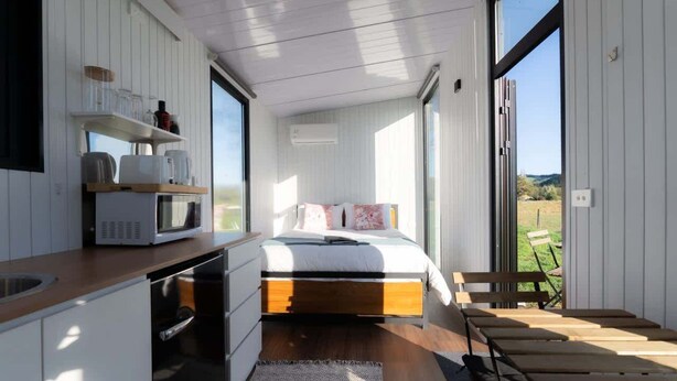 Un dormitorio moderno con cama, mesitas de noche, microondas integrado y un ventanal grande.