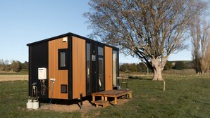 Una cabaña pequeña y moderna con fachada de madera y ventanas amplias, ubicada en un campo cubierto de hierba con un árbol cercano.