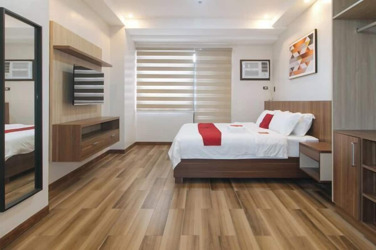 Une chambre d’hôtel moderne avec un plancher en bois, un lit avec une literie blanche et des touches de couleur rouge, un téléviseur fixé au mur et une fenêtre avec des stores.