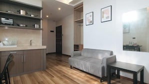 A modern living room with a grey sofa, a small table, and a kitchen area with wooden flooring and built-in shelves. - RedDoorz Plus @ Bez Tower and Residences San Juan (San Juan)