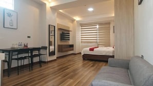 A modern hotel room with a wooden floor, a bed with a red accent, a grey sofa, a dining area with chairs, and a wall-mounted TV. - RedDoorz Plus @ Bez Tower and Residences San Juan (San Juan)
