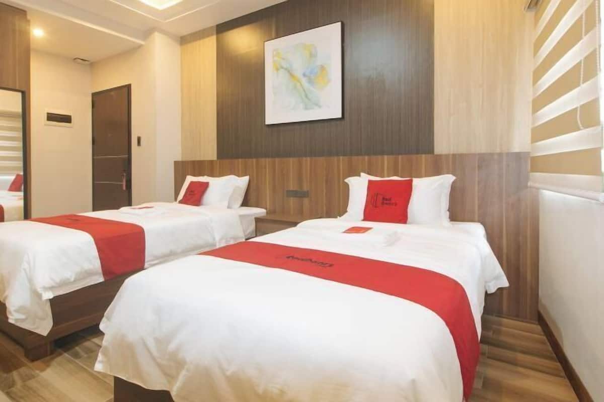 Une chambre d’hôtel avec deux lits, chacun agrémenté de draps blancs et de touches de couleur rouge, une tête de lit en bois, un tableau abstrait encadré et un plancher en bois.
