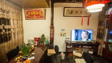A traditional room with a wooden ceiling, a television, a desk with a computer, and a dining table with fruits and a vase.