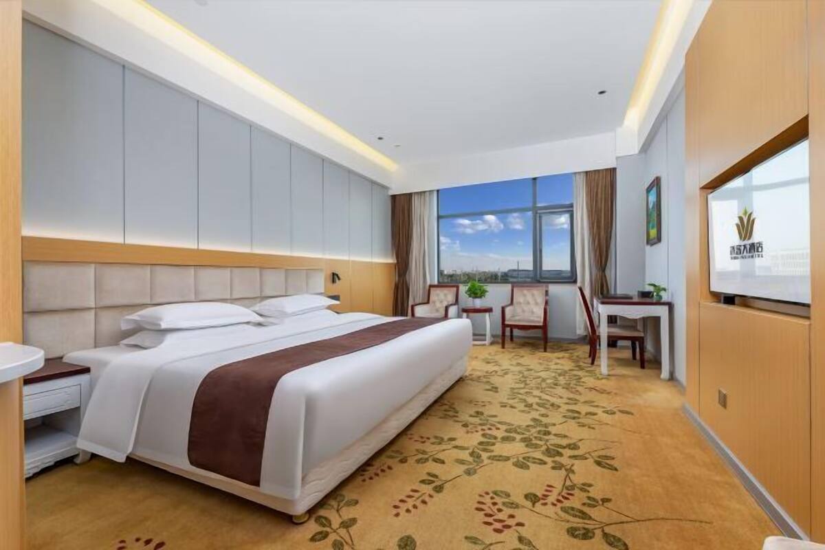A hotel room with a large bed, a desk, a chair, a small table, a window with a view, and a television on the wall.
