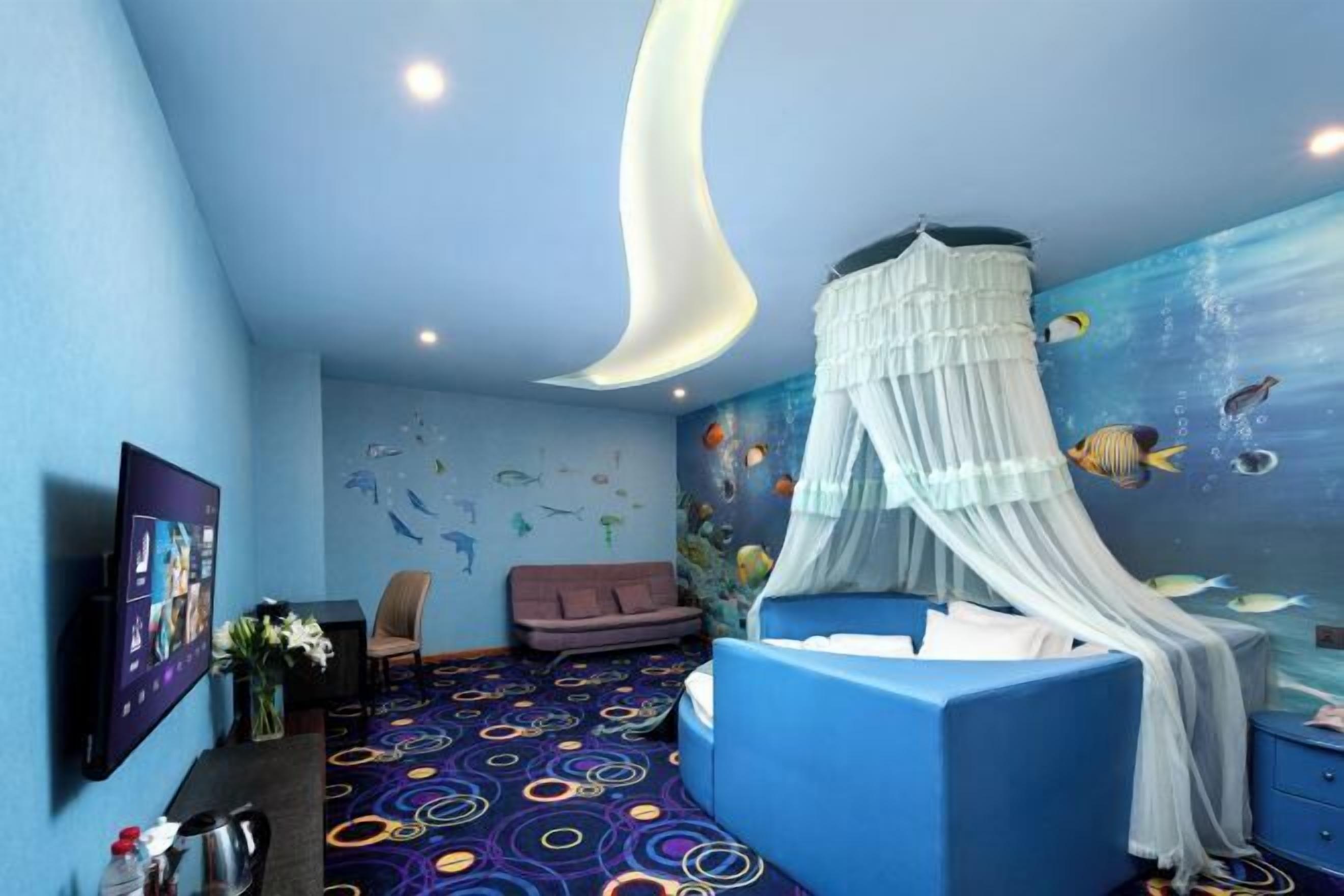 A room with a bed, a TV, a sofa, and a wall mural of fish.