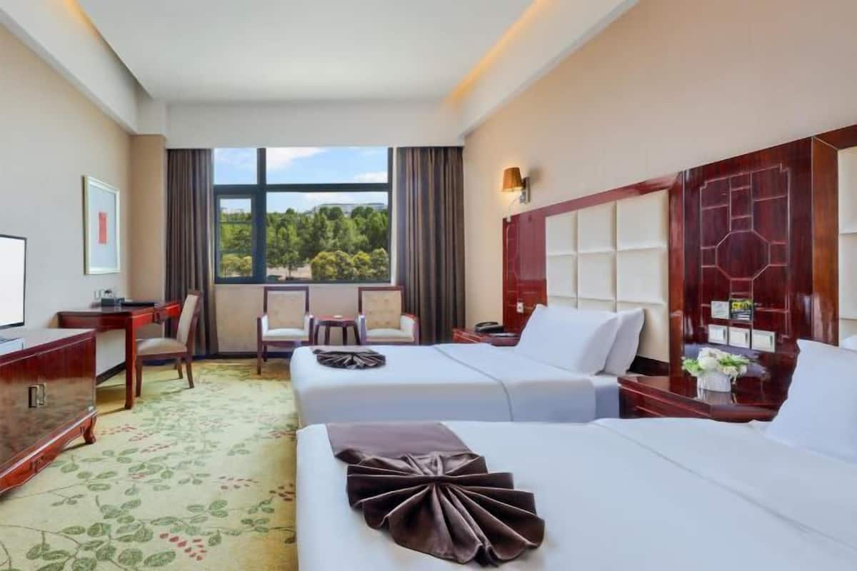 A hotel room with two beds, a desk, a chair, a TV, and a large window with a view of trees.
