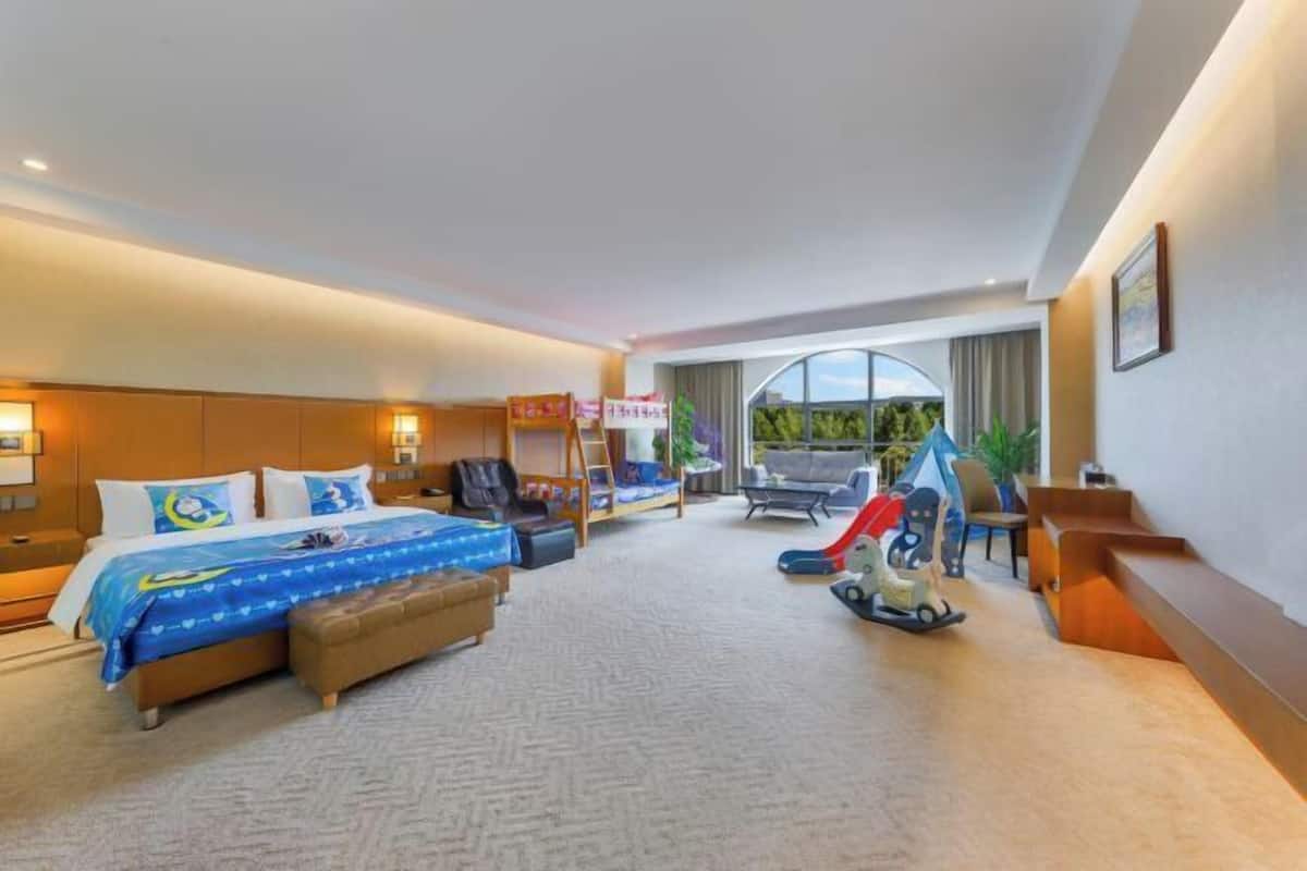 A hotel room with a bed, a desk, a chair, a small table, a play area with toys, and a window with a view.