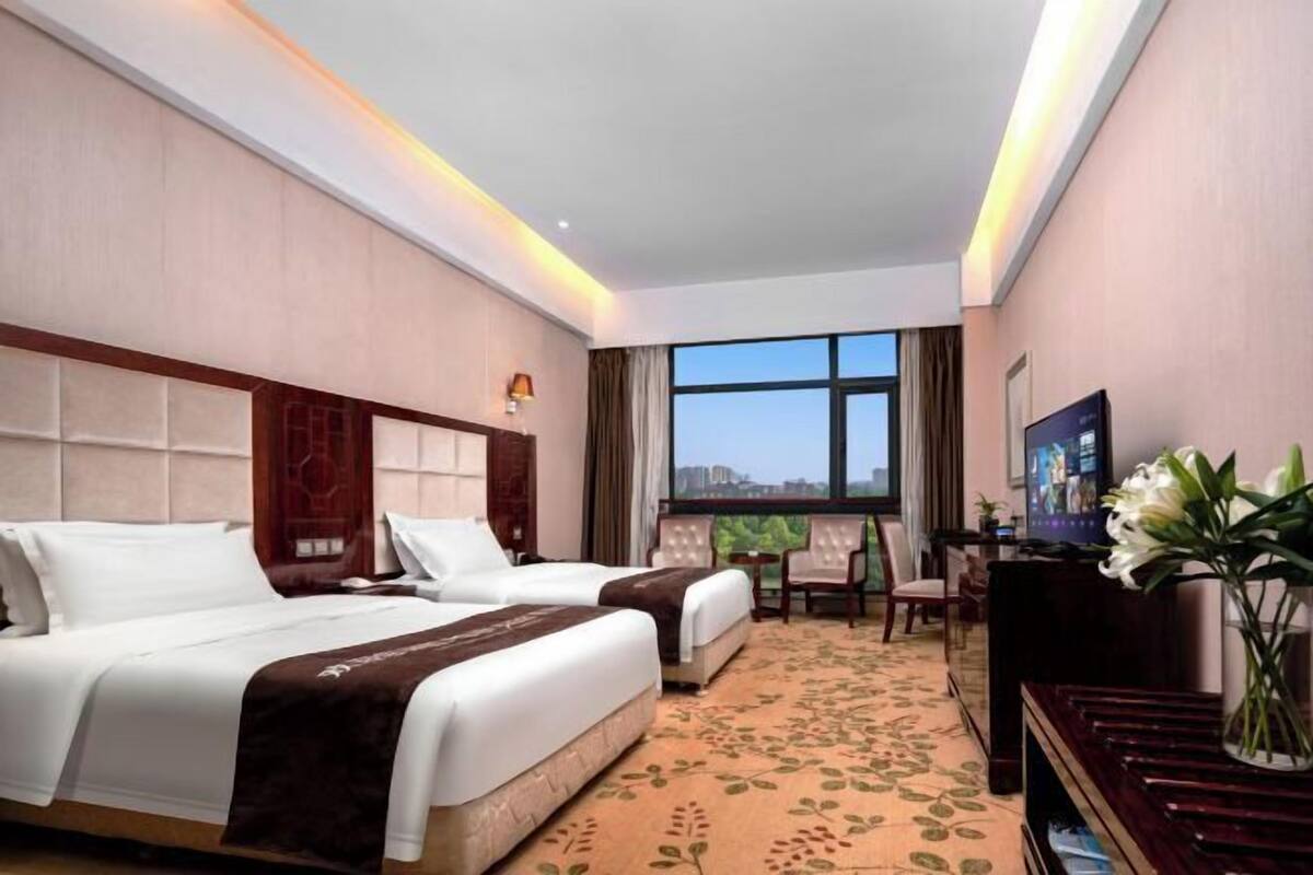 A hotel room with two beds, a desk, a chair, a TV, and a large window with a city view.